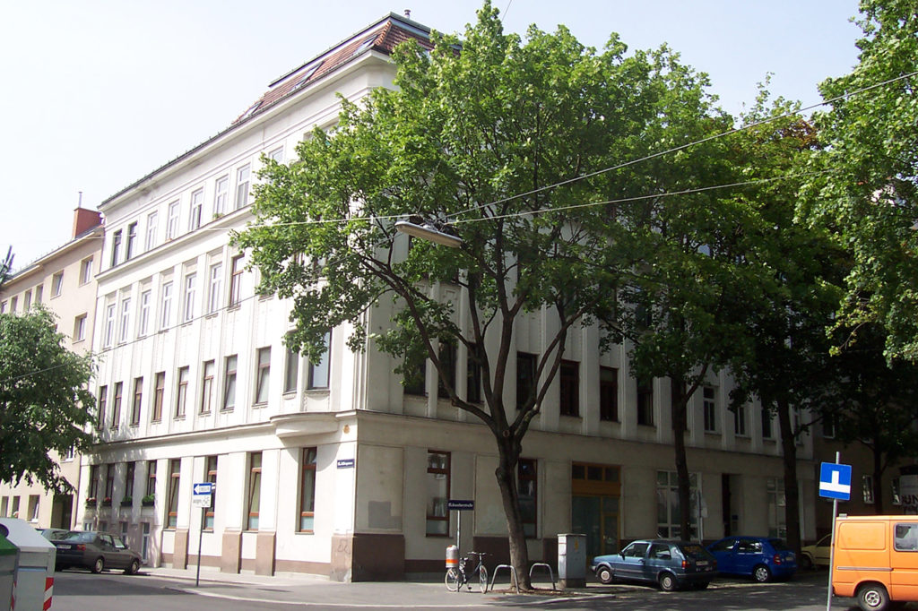 Eckzinshaus im 15. Bezirk Wiens, weiße Fassade, davor große grüne Laubbäume