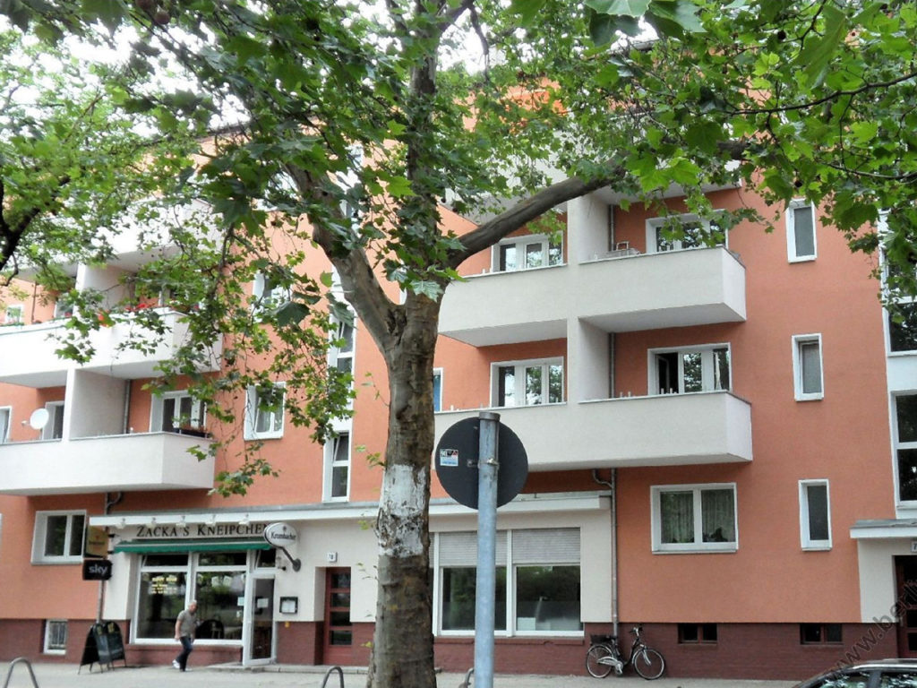Wohnhausanlage in Berlin, Geschäftslokal im Erdgeschoß, großer Laubbaum im Vordergrund, weiße Balkone und Fenster, Fassade lachsfarben