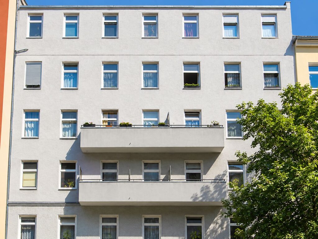 Zinshaus mit hellgrauer Fassade, Balkone auf zwei Stockwerken, rechts davor ein Baum