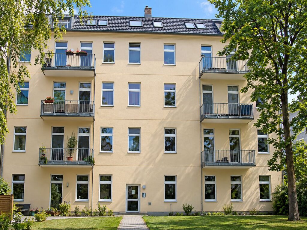 Wohnhaus mit cremfarbiger Fassade, Balkone, links und rechts davor ein Baum